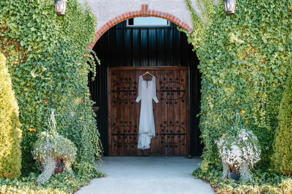 Long sleeve bridal gown hanging in archway of venue. Archway is covered with green moss on either side.