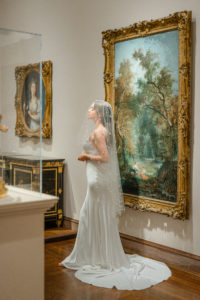 Bridal portrait in the Cincinnati Museum of Art. Bride is wearing a fitted gown with train and a pearled veil over her face in front of paintings