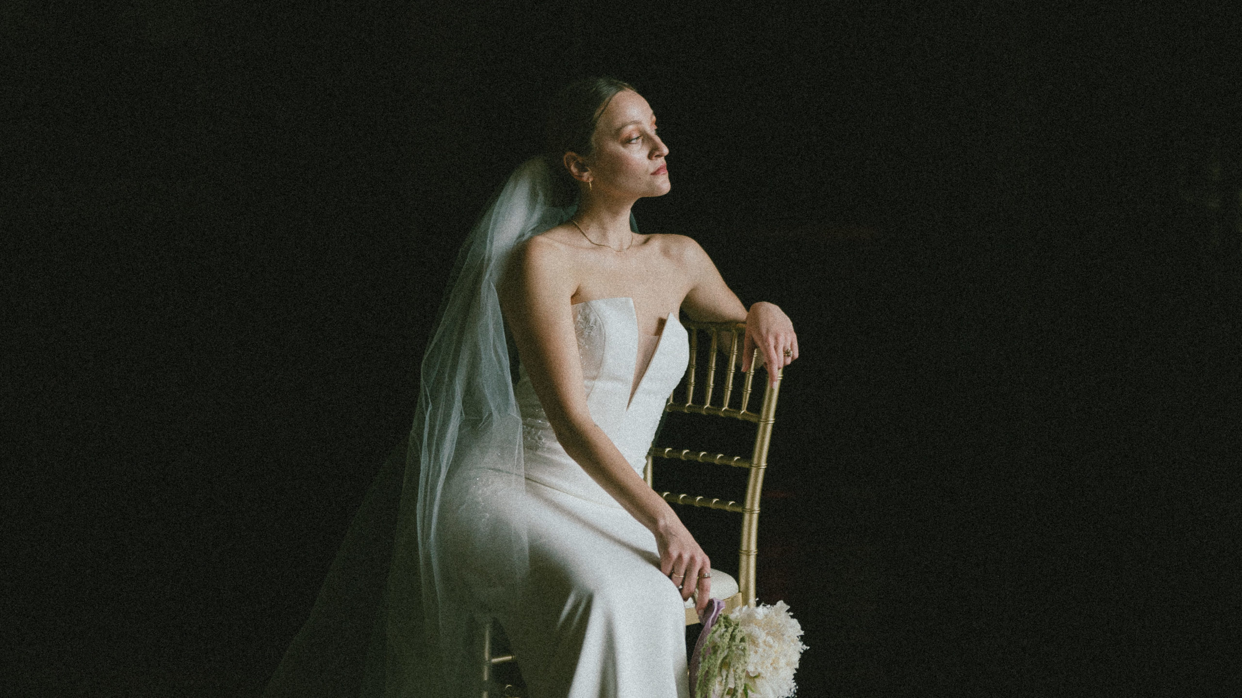 Bridal portrait of a woman sitting in a chair looking off into the distance wearing a strapless gown and a long veil
