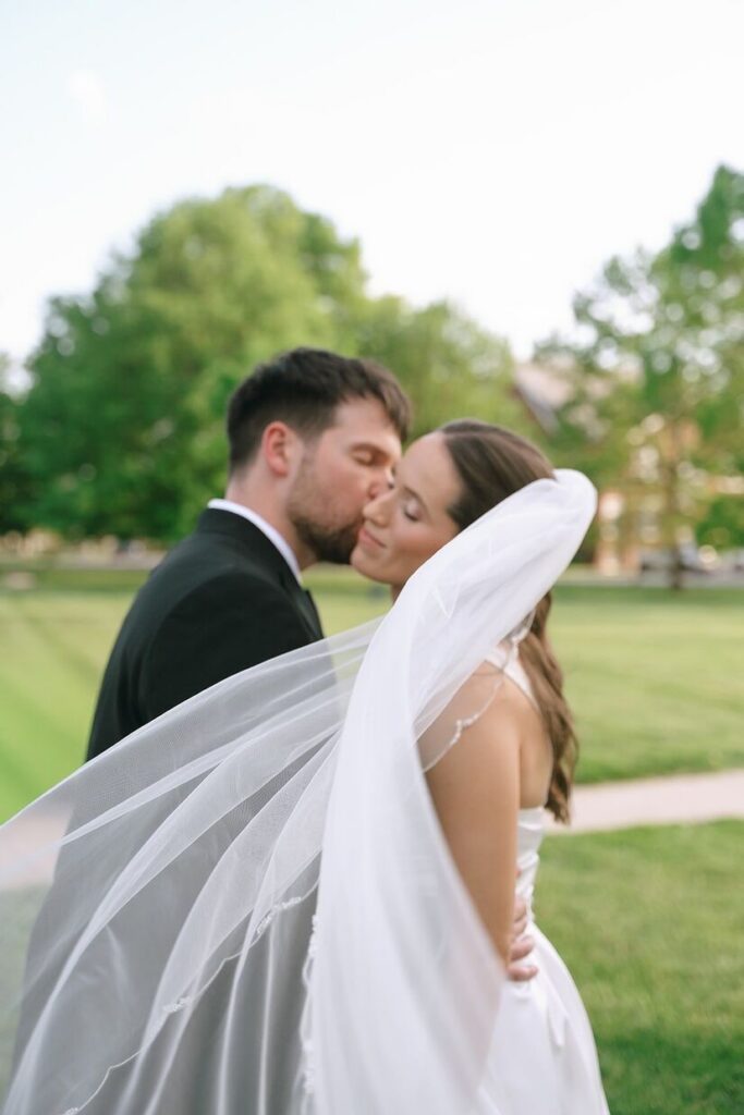 Groom kissing bride with her veil blowing in the wind at The Olmstead, a venue for outdoor weddings in Louisville. 