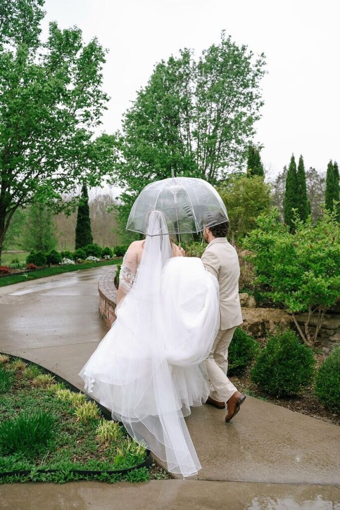 Groom holding umbrella over bride as they walk around the gardens at Hazelnut Farms, a venue for outdoor weddings in Louisville. 