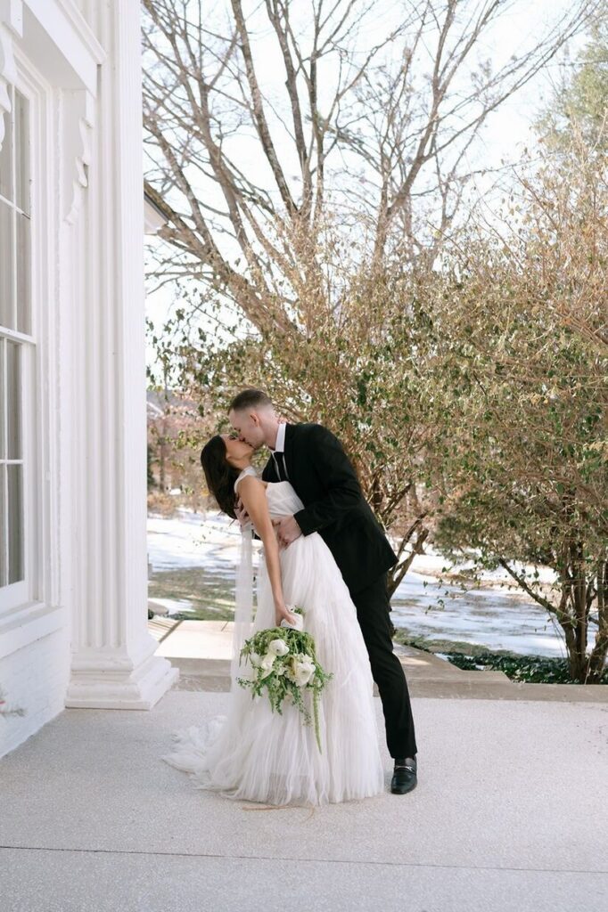 Groom dipping and kissing bride in a snowy landscape with an elegant building next to them at Whitehall, a venue for outdoor weddings in Louisville.