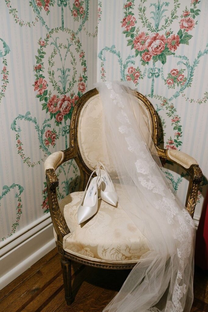 A vintage chair sitting against a wall with floral wallpaper and a pair of heels on it for a Whitehall mansion wedding. 